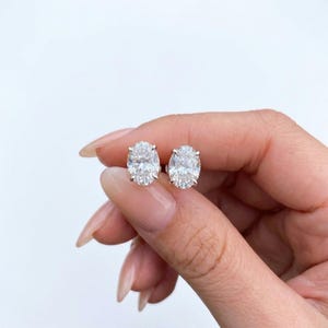 Puede incluir: Un par de pendientes de botón con piedras transparentes de corte ovalado montadas en garras de metal plateado. Cada pendiente muestra una superficie facetada que refleja la luz, exhibiendo un aspecto brillante y transparente con cuatro garras.