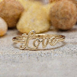 May include: A gold-toned ring featuring the word Love in a cursive script font. The ring has a polished metallic finish and a thin band that connects to the lettering. The design is centered and clearly legible, showcasing the script style of the word.