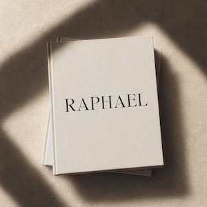 May include: A stack of three hardcover books with off-white covers. The word "RAPHAEL" is printed in black, centered on the top book. The books are on a textured, light beige surface, with shadows across the scene.