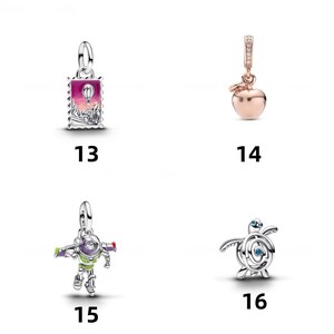 May include: Four silver and rose-gold tone charms labeled 13 through 16. Charm 13 is a rectangular stamp with a hot air balloon. Charm 14 is a rose-gold apple. Charm 15 is a colorful space ranger figure. Charm 16 is a silver sea turtle charm.