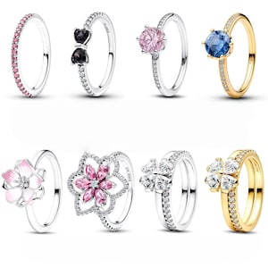 May include: Collection of silver and gold rings with various designs. Rings include a pink gemstone band, a bow design with black stones, a pink gemstone solitaire, and a blue gemstone solitaire. Other rings feature floral and clover designs with white and pink gemstones.