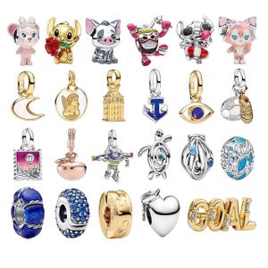 May include: A collection of twenty-four metal charms featuring various designs including cartoon characters, a crescent moon, an angel, a building, an anchor, an eye, a soccer ball, a shoe, a stamp, a fruit, a space ranger, a turtle, and the word GOAL.