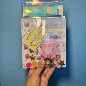 May include: A clear cellophane package containing craft supplies. The package includes decorative paper, stickers, a small pink bag labeled "TICKET", and a yellow tag with the text "Dulce Crafts". The background is a light blue.