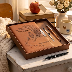 Puede incluir: Un diario con cubierta de cuero color canela y dos bolígrafos presentados en una caja de regalo marrón. La cubierta del diario presenta una ilustración grabada con láser de dos osos caminando entre árboles con el texto: Never forget the difference you make Mrs. Sophia Thank you, Kim.