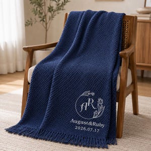 May include: A navy blue textured throw blanket with fringe edges, featuring white embroidery of the initials AR inside a floral wreath, followed by the text August & Ruby and the date 2026.07.17, draped over a wooden chair with cane accents.