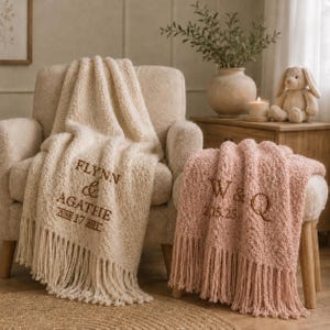 May include: A cream-coloured textured throw blanket with fringe, embroidered with the text FLYNN & AGATHE JUNE 17 2025, and a pink textured throw blanket with fringe, embroidered with the text W & Q 2.15.25, both draped over furniture.