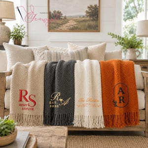 May include: Four textured knit throw blankets with fringe, each featuring custom embroidery. The beige blanket has red 'RS' and 'REMY & SYLVIE' text. The charcoal blanket has gold 'RA' and 'EST. 2026'. The cream blanket has gold script text. The orange blanket has a black 'AR' monogram.