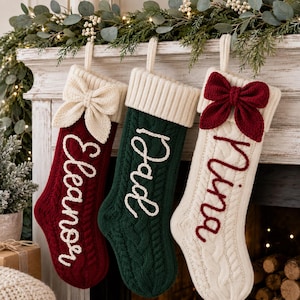 May include: Three cable-knit Christmas stockings hanging in a row. The left is red with a cream bow and the name Eleanor. The center is dark green with a cream cuff and the name Dad. The right is cream with a red bow and the name Nina.