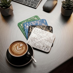 Könnte beinhalten: Vier quadratische Untersetzer mit Leiterplattendesigns in Grün, Blau, Schwarz und Silber. Eine Tasse Kaffee mit Latte Art steht auf einer schwarzen Untertasse. Eine Tastatur, eine Maus und Sukkulenten sind ebenfalls zu sehen.