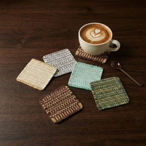 May include: A set of five square woven coasters in beige, grey, light green, dark green, and brown, accompanied by a white ceramic mug filled with coffee featuring latte art, a matching coaster underneath the mug, and a small brown spoon.