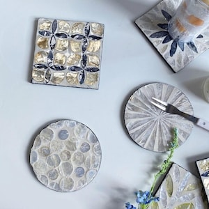 May include: A collection of decorative coasters with mosaic tile designs. The coasters feature a variety of patterns, including floral and circular designs, with gold, silver, and blue accents. One coaster has a fork on it.