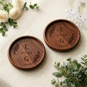 May include: Two round, wooden coasters with engraved initials and date. Each coaster features the letters "L" and "G" with the date "05.26.2026" and floral accents. The coasters are a dark brown color and are set on a light-colored surface.