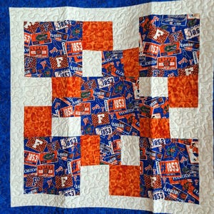 May include: A handmade quilt featuring a patchwork design with a blue, orange, and white color scheme. The quilt showcases a repeating pattern of squares with the text "FLORIDA" and "1853" printed on the fabric. The quilt has a blue border.
