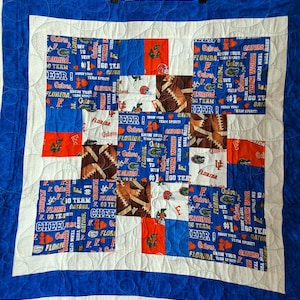 May include: A handmade quilt featuring a patchwork design with blue, orange, and white squares. The squares display footballs, the word "Gators", and team spirit phrases. The quilt has a blue border and is likely a fan item.