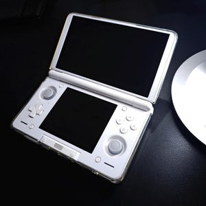 May include: A white handheld gaming console with a dual-screen clamshell design. The device features a top screen, a bottom touchscreen, a directional pad, four action buttons labeled A, B, X, and Y, two analog sticks, and a clear protective case.