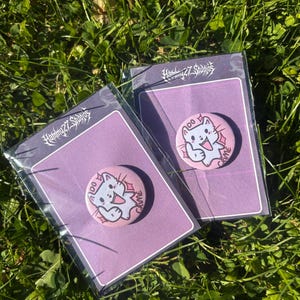 Puede incluir: Dos pines redondos con un diseño de gato de dibujos animados sobre una tarjeta de respaldo morada. El gato es azul claro con acentos rosas y el texto "do crime". Las tarjetas están empaquetadas en plástico transparente, sobre hierba verde.
