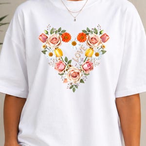 Può includere: T-shirt bianco con un motivo floreale a forma di cuore. Il cuore è composto da rose rosa, tulipani gialli, gerbere arancioni e margherite bianche, con foglie verdi. È visibile una collana d'oro.