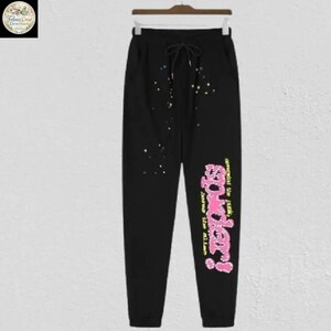 May include: Black sweatpants with a drawstring waist and colorful splatter paint design. The right leg features the text "Spider!" in pink and yellow, with additional text below. The pants are hanging on a wooden hanger.