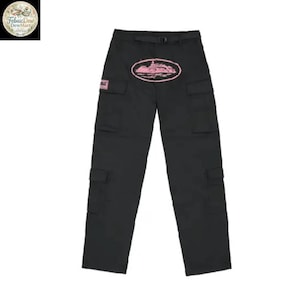 May include: Black cargo pants with multiple pockets and a belt. A pink oval graphic with a building design is centered on the front. A small pink square with text is on the left pocket.