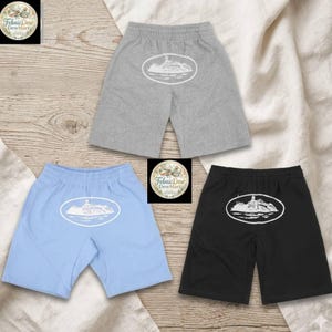Puede incluir: Tres pares de pantalones cortos en gris, azul claro y negro. Cada par presenta un dise&ntilde;o ovalado blanco con una ilustraci&oacute;n detallada de un edificio. Los pantalones cortos tienen cintura el&aacute;stica y un estilo casual.