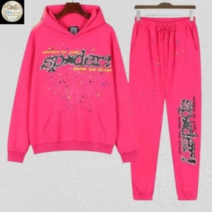 May include: A bright pink hoodie and matching sweatpants set. The hoodie features a front pocket and a graphic that reads "Spider" in a stylized font. The sweatpants have a drawstring waist and a matching graphic on the leg. The set is decorated with colorful paint splatters.