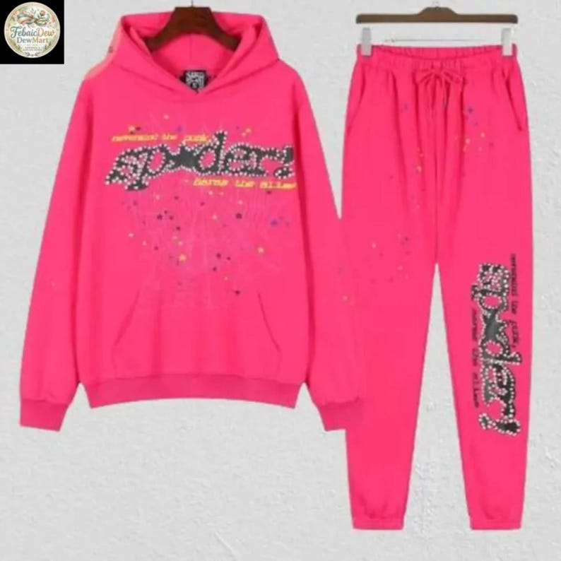 May include: A bright pink hoodie and matching sweatpants set. The hoodie features a front pocket and a graphic that reads "Spider" in a stylized font. The sweatpants have a drawstring waist and a matching graphic on the leg. The set is decorated with colorful paint splatters.