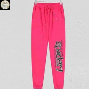 May include: Bright pink sweatpants with a drawstring waist and pockets. The pants feature a decorative design on the left leg with the word "Doodle" in a stylized font. Small, colorful paint splatters are scattered across the front.
