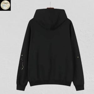 May include: A black hooded sweatshirt with a drawstring hood. The back of the sweatshirt features a simple design of gold-colored dots on the left sleeve and a single dot on the right sleeve. The sweatshirt is hanging on a hanger.