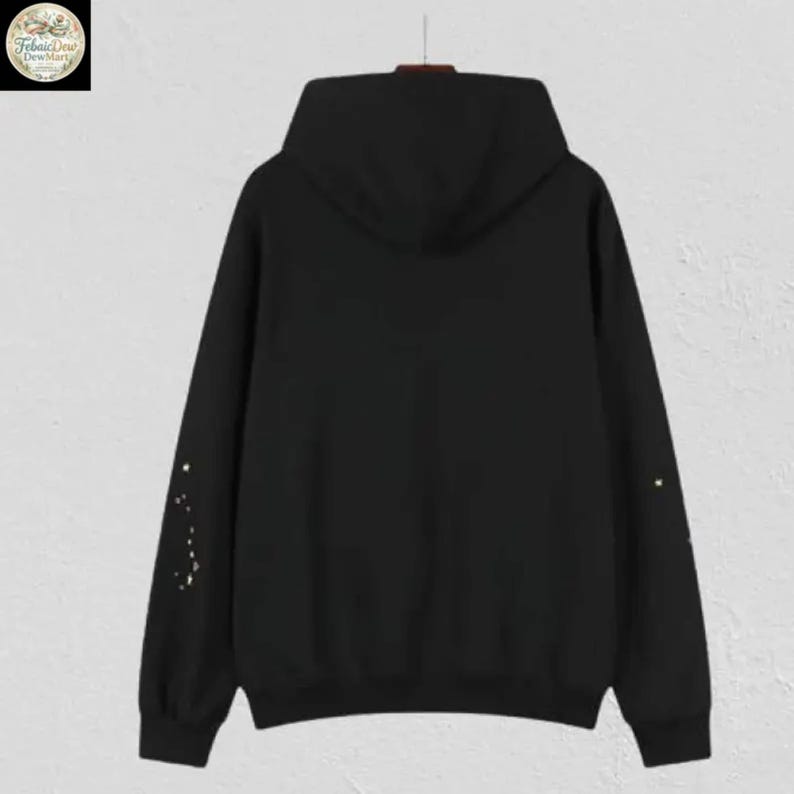 May include: A black hooded sweatshirt with a drawstring hood. The back of the sweatshirt features a simple design of gold-colored dots on the left sleeve and a single dot on the right sleeve. The sweatshirt is hanging on a hanger.