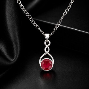 May include: A silver-toned necklace featuring a pendant with a twisted metal design at the top and a round, faceted deep red gemstone at the bottom. The pendant hangs from a delicate silver-toned chain with circular links, creating a simple look.