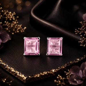 Puede incluir: Un par de pendientes de botón con gemas rectangulares de color rosa claro en talla esmeralda, montadas en garras de metal plateado. Los pendientes están colocados uno al lado del otro, mostrando las superficies facetadas de las piedras y la calidad transparente del material rosa sobre una superficie oscura y texturizada.