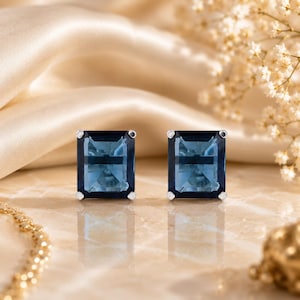 May include: A pair of stud earrings featuring rectangular, emerald-cut blue gemstones set in silver-toned metal prongs. The stones exhibit a deep, translucent blue hue with faceted surfaces that reflect light, creating a clear, polished appearance.