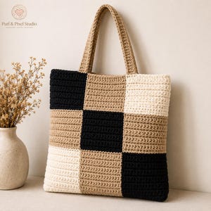 Puede incluir: Un bolso tote cuadrado de ganchillo con un patrón de cuadros en hilo negro, beige y crema. El bolso tiene dos asas de ganchillo beige a juego y una apariencia texturizada cosida a mano. El diseño consiste en cuadrados de colores alternos.