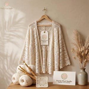 Op de afbeelding: Een gebroken wit gehaakt vest met een open voorkant en wijde mouwen, hangend aan een houten kledinghanger. Een label met de tekst Purl & Pixel Studio is bevestigd. Daaronder liggen verschillende bollen garen, een haaknaald en een gehaakt vierkantje.