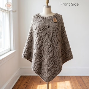 Könnte beinhalten: Ein taupefarbener Strickponcho mit einem markanten Zopfmuster in Blattform auf der Vorderseite, einem gerippten Rundhalsausschnitt und einem gerippten Saum. Das Kleidungsstück wird auf einer Schneiderpuppe präsentiert und hat ein kleines braunes Etikett.