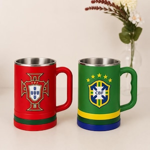Pode incluir: Duas canecas cilíndricas de metal com alças. A caneca da esquerda é vermelha com base verde e apresenta o escudo da FPF de Portugal. A caneca da direita é verde com base amarela e azul e apresenta o escudo da CBF do Brasil com cinco estrelas.