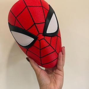 Könnte beinhalten: Eine rote Spider-Man-Maske mit einem schwarzen Spinnennetz-Muster auf der Oberfläche. Die Maske verfügt über zwei große, weiße, netzartige Augenlinsen, die von dicken schwarzen Rändern umrahmt sind und ein markantes Aussehen verleihen.