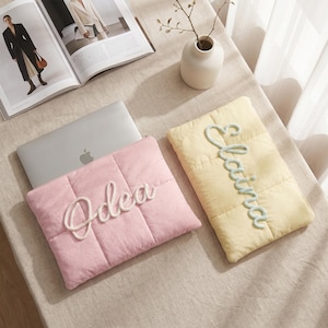 May include: Two rectangular quilted laptop sleeves, one in soft pink and one in pale yellow. The pink sleeve features the name Odea in white cord lettering, while the yellow sleeve displays the name Elaina in light green cord lettering.