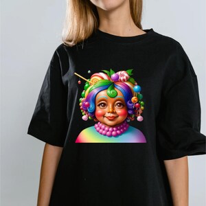 May include: A black short-sleeved t-shirt featuring a graphic print of a character with vibrant, multi-colored hair styled with various round candies and lollipops. The character wears a pink beaded necklace and a rainbow-colored shirt collar.