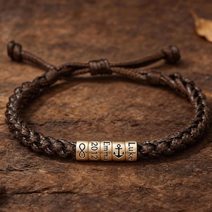 May include: A dark brown braided cord bracelet featuring five cylindrical metal beads. The beads are engraved with an infinity symbol, the year 2012, the name Emma, an anchor icon, and the name Luke. The bracelet has adjustable sliding knots.