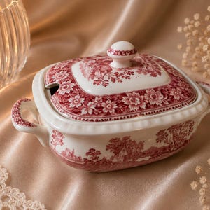 Könnte beinhalten: Eine weiße Keramik-Terrine mit einem roten Transferdruck-Muster aus Blumenmotiven und einer Landschaftsszene. Das ovale Gefäß verfügt über zwei seitliche Griffe, einen passenden Deckel mit Knauf und eine Aussparung für einen Servierlöffel.