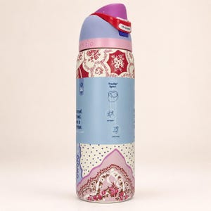 May include: A cylindrical Owala water bottle featuring a light blue and cream base with a red and white floral pattern. The lid is light purple with a red locking mechanism labeled Lift to Unlock. Text on the bottle details the FreeSip spout features.