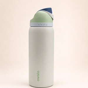 Pu&ograve; includere: Una borraccia Owala grigio chiaro con un coperchio verde pallido e blu scuro. La bottiglia presenta un corpo cilindrico con il nome del marchio Owala stampato in verde vicino alla base e in rilievo bianco sulla parte grigia del coperchio.