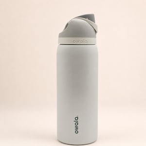 Pu&ograve; includere: Una borraccia cilindrica grigio chiaro con finitura opaca. La borraccia presenta un tappo grigio scuro e bianco con un manico integrato per il trasporto. Il marchio Owala &egrave; stampato in grigio scuro sul corpo e impresso sul colletto del tappo.