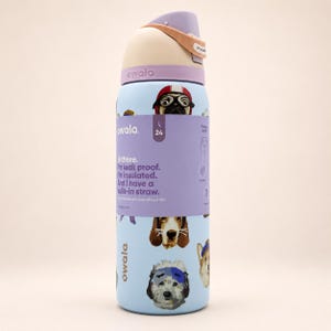 May include: A light blue Owala water bottle with a 24-ounce capacity, featuring a wrap-around label with text and dog illustrations. The bottle has a lavender and beige lid with a built-in straw, and the label reads: Hi there. I'm leak proof.