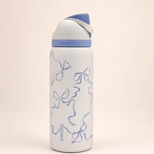 May include: A white Owala water bottle featuring a light blue bow pattern printed on the body. The bottle has a light blue and white lid with a carry loop and the brand name Owala printed in light blue on the base and the lid collar.