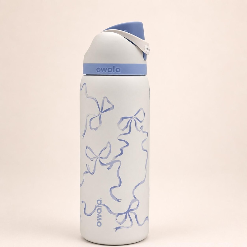 May include: A white Owala water bottle featuring a light blue bow pattern printed on the body. The bottle has a light blue and white lid with a carry loop and the brand name Owala printed in light blue on the base and the lid collar.