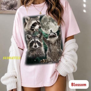 Könnte beinhalten: Ein hellrosa T-Shirt mit kurzen Ärmeln und einem Grafikdruck von drei Waschbären und einem Vollmond. Ein Waschbär streckt sich zum Mond, während zwei weitere darunter abgebildet sind. Der Text SUSMIATEES ist in Gelb auf der linken Seite.