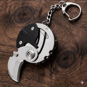 Op de afbeelding: Een compact vouwmes van roestvrij staal met een zwarte handgreep, bevestigd aan een ketting en sleutelhanger. Het lemmet is gebogen en open. Het mes wordt getoond op een donker houten oppervlak.