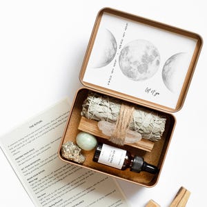 May include: A rectangular copper-colored metal tin containing a sage bundle, a palo santo stick, a small clear quartz crystal, a smooth green stone, a pyrite cluster, and a dark glass dropper bottle labeled HEALING. The lid features moon phases.
